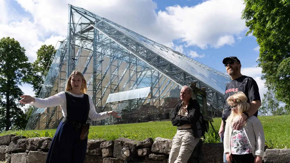 Guide i middelalderdrakt forteller med ivrige gester om Domkirkeoddens historie til to menn og et barn. Hamardomen i bakgrunnen.