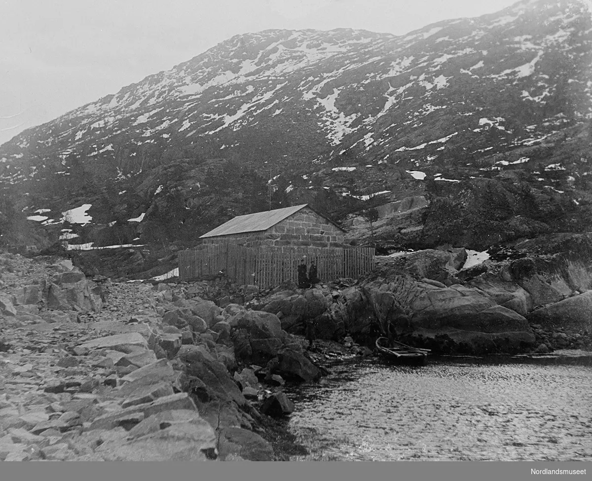Narvik, dynamittlager i Indre Sildvik. Bygning av stein med plankegjerde rundt. Bølgeblikk på taket. Fjell i bakgrunnen og litt av havet til høyre.