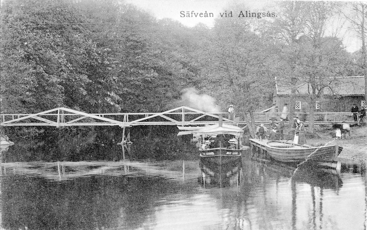 Vid bron som sträcker sig över Säveån ses en fullsatt ångslup "Jonas Alströmer" en större eka samt en smärre folksamling vid byggan. Text på kortet "Säfveån vid Alingsås"
