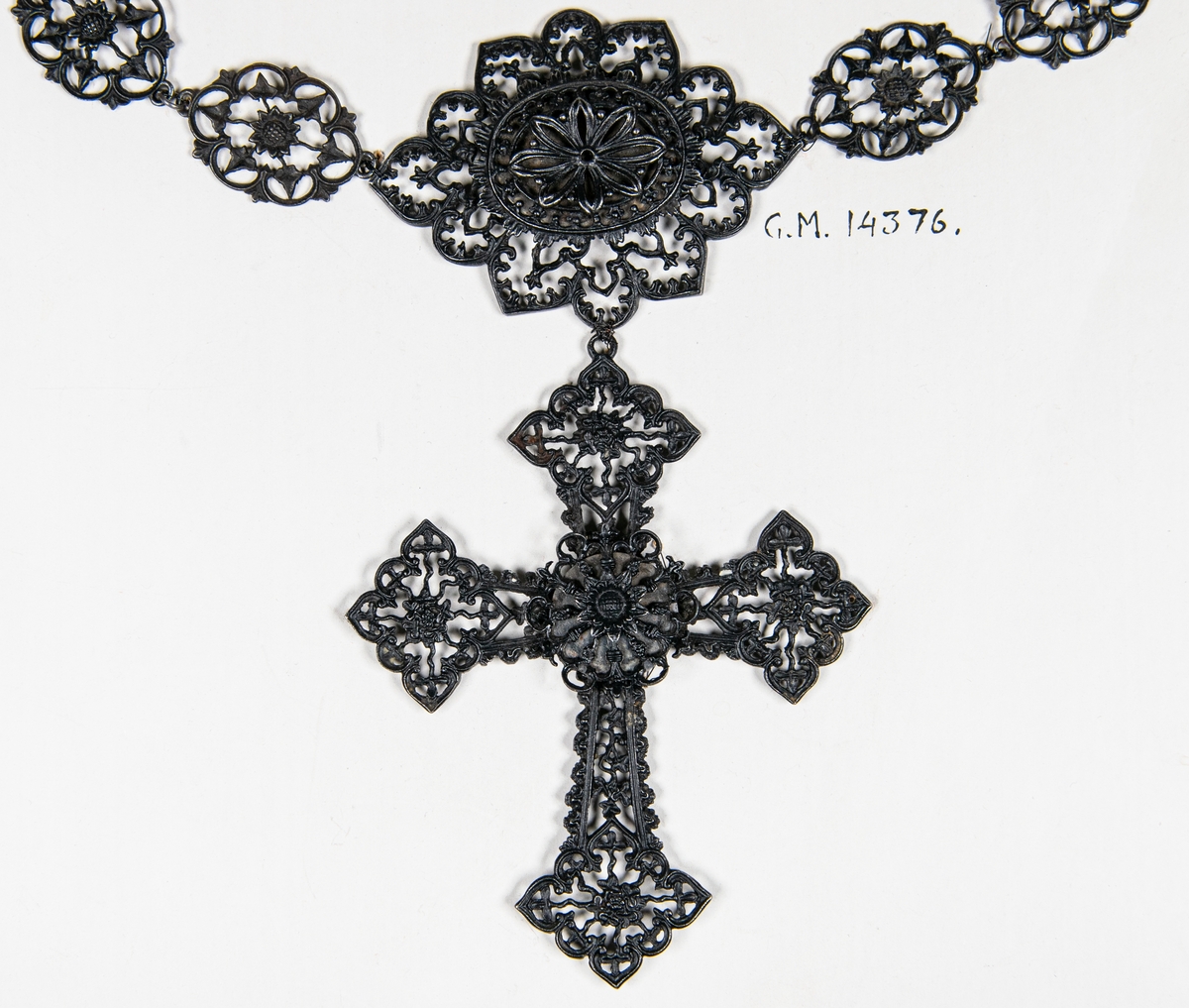 Smycke, halsband av metall, gjutet, genombrutna plattor, i mitten en större varvid ett kors är fastsatt. Lås: två händer som fattar varandra.