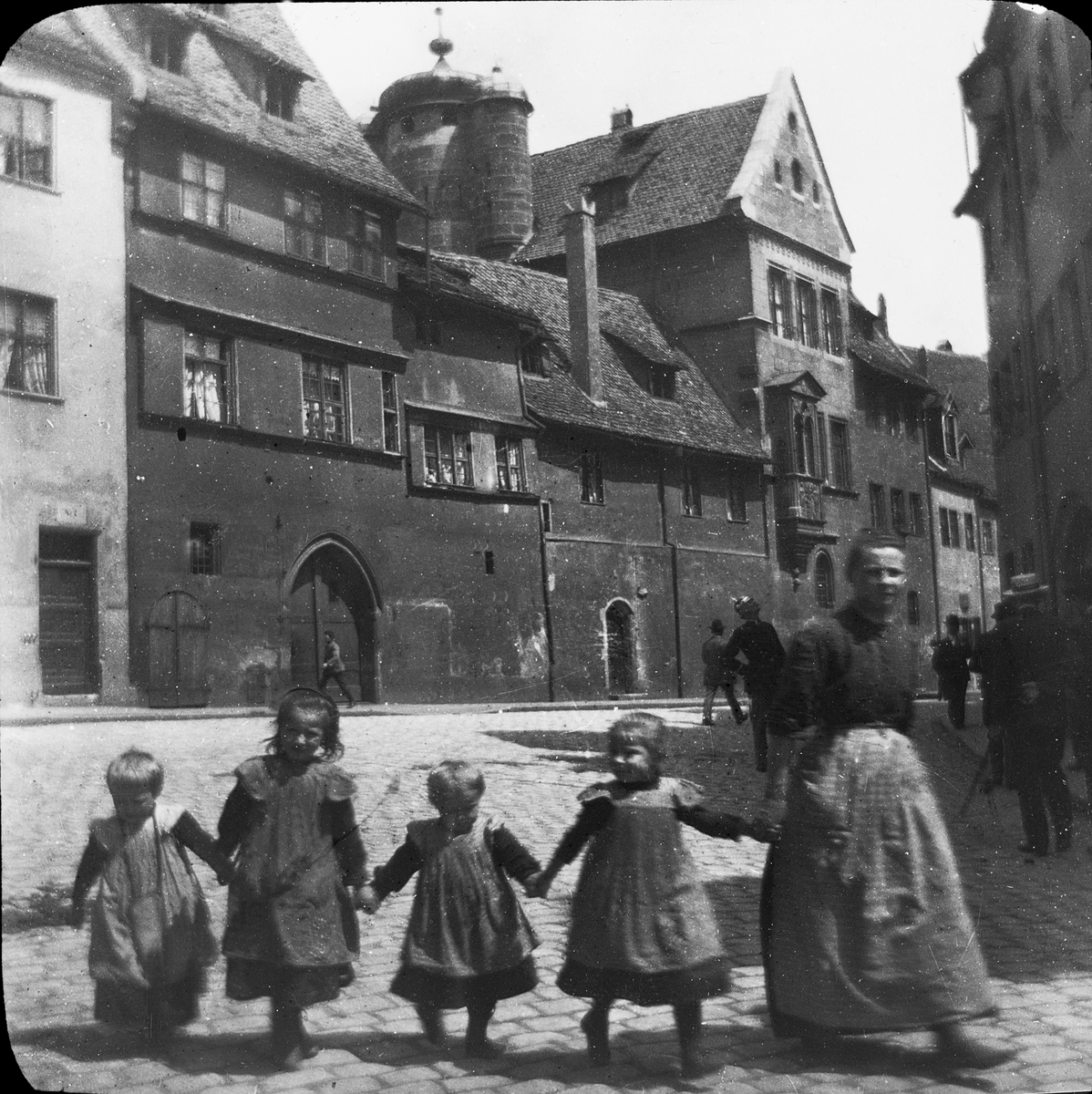 Skioptikonbild med motiv av kvinna med fyra små barn på gata i Nürnberg.
Bilden har förvarats i kartong märkt: Nürnberg VI. 1901