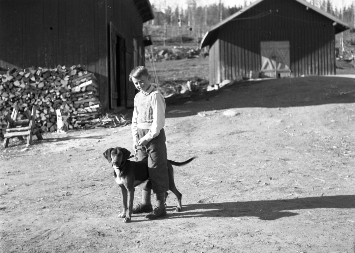 En gutt med en hund stående på gårdsplass. Vedstabel og bygninger sees i bakgrunnen.
