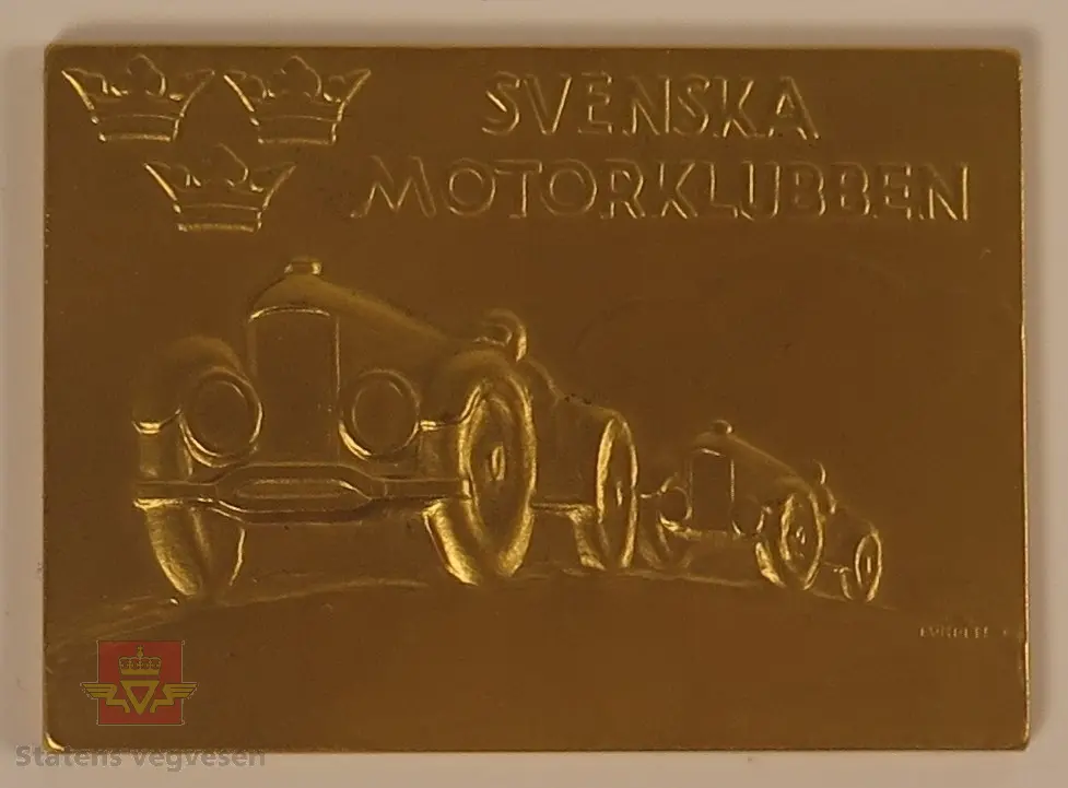 Gruppering av plaketter laget av metall i gull farge. På plakettene er det utformet to biler som tilsynelatende kjører om kapp. Det er også utformet tre kongelige kroner.