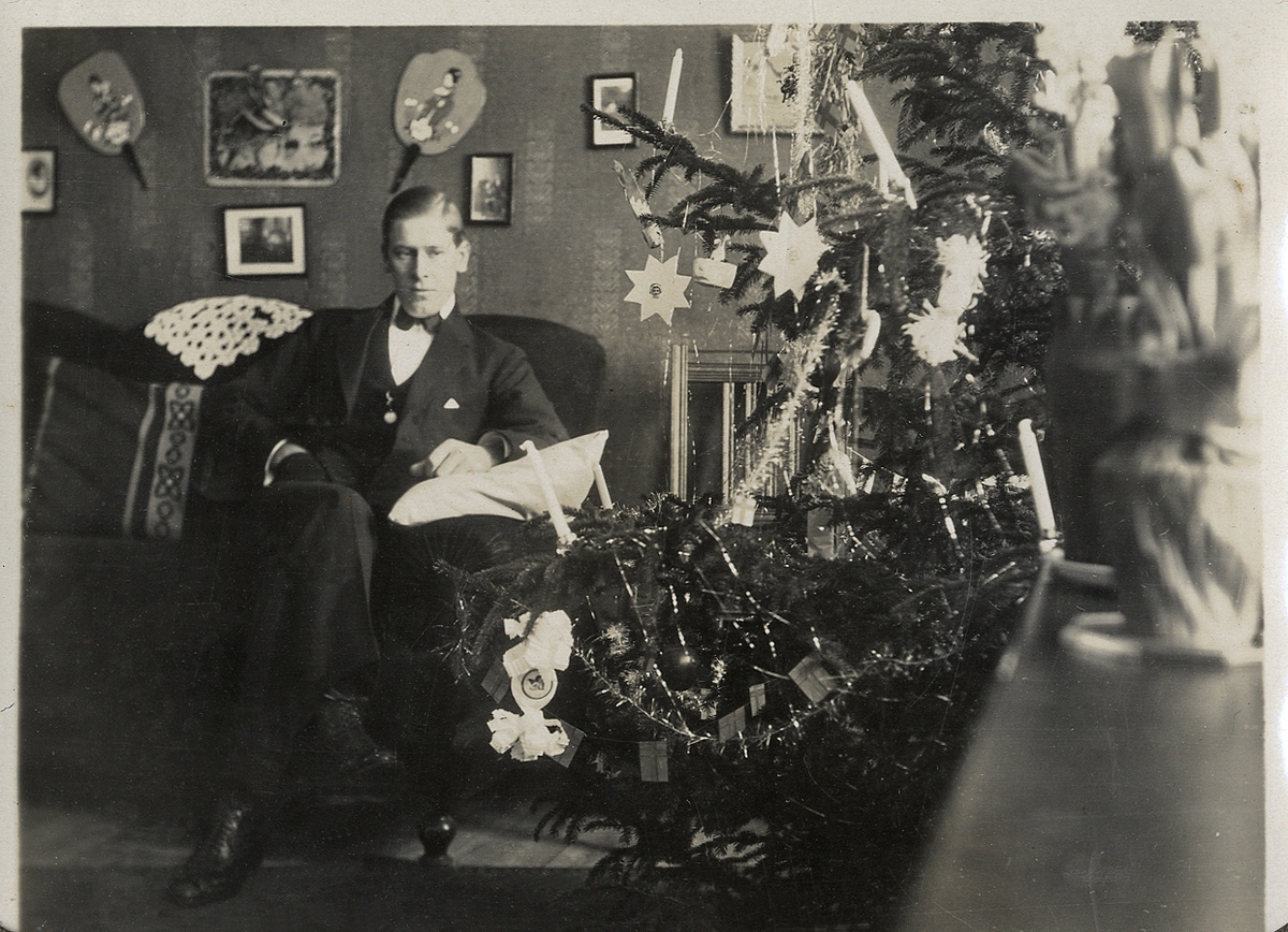 En man i kostym sitter i en soffa i ett vardagsrum. Till höger syns en julgran.