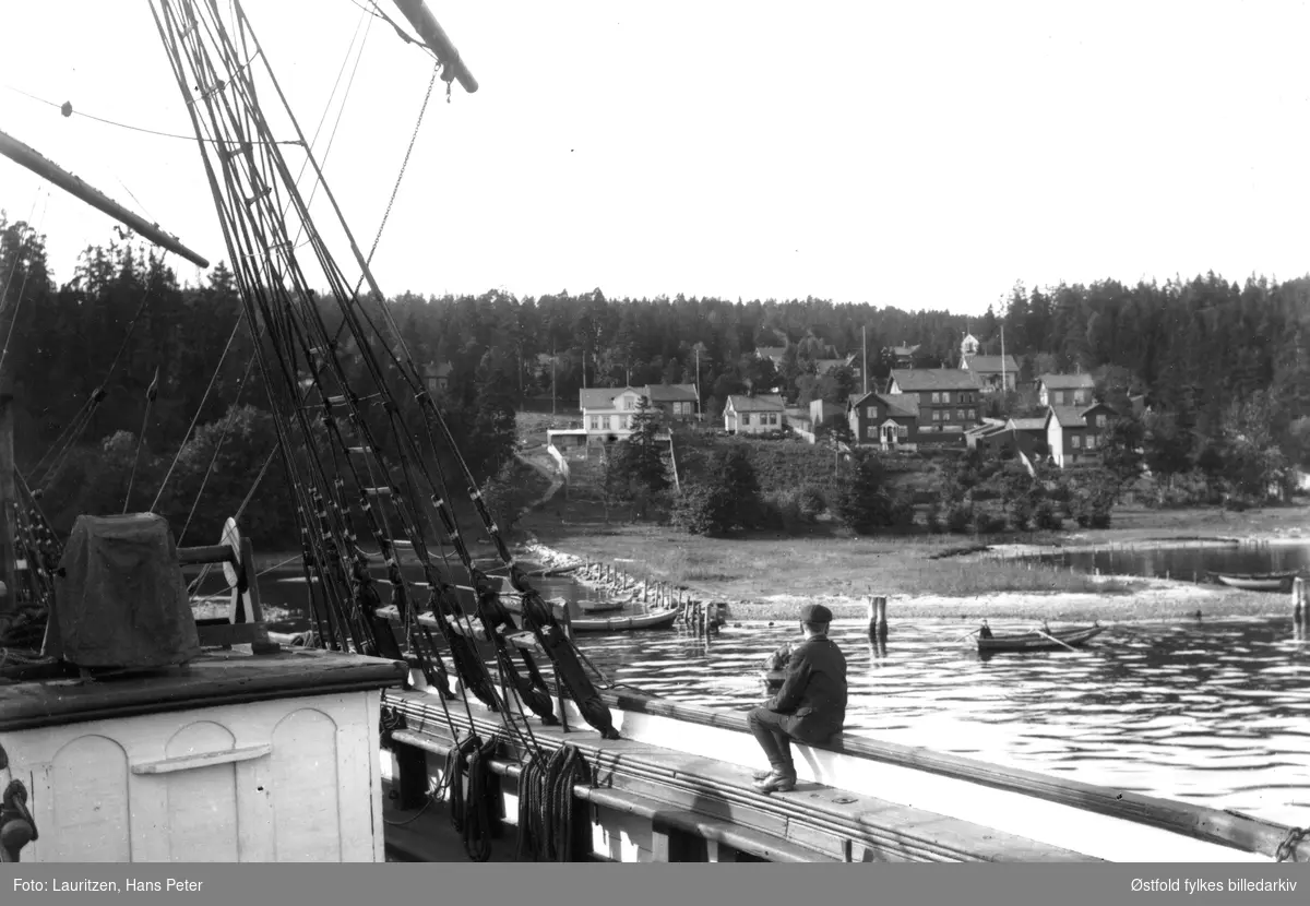 Gutt sitter på rekka av en seilbåt. Flere robåter, boligområde i bakgrunnen. "Betongen - Jeløy" (?)