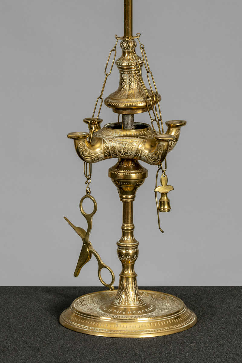 Oljelampe/ romersk lampe i messing med 4 armer/vekebrennere. Påmontert 4 kjettinger, hvorav en holder vekesaks, en slokker og to messingnåler (?) Har tilhørt Sara Fabricius.