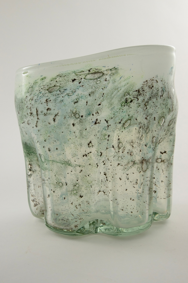 Irregulær sylinderformet vase i halvgjennomskinnelig melkehvitt glass. Ujevn munningsrand. Korpus er derkorert med metallspon, luftblærer og blå-, sort- og grønnfargede partier.