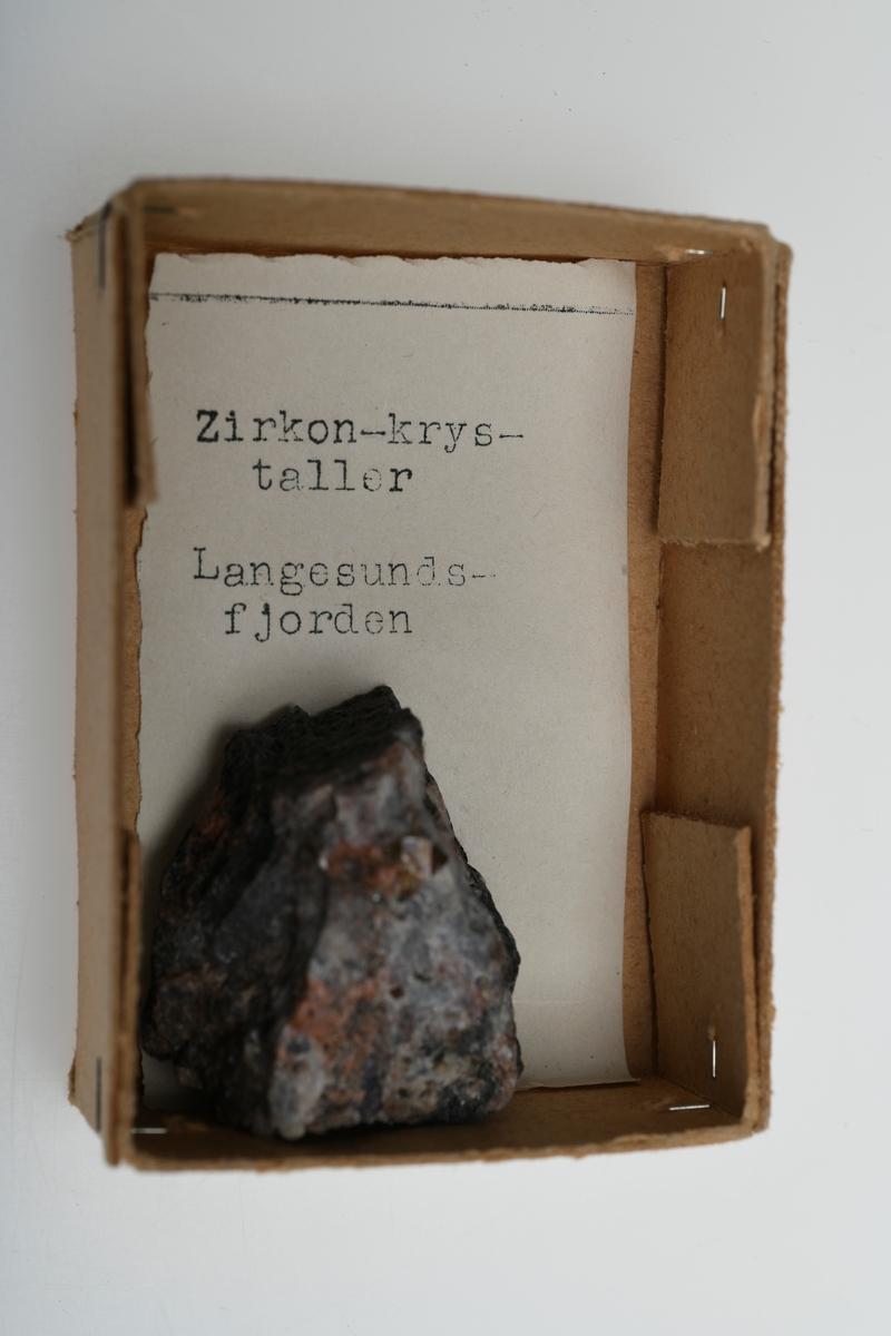 Et stykke zirkon (silikatmineral) fra Langesundsfjorden, Telemark. Fargen er grå med innslag av rødt og brunt samt hvitt. Zirkonen kan sees som en liten blank og brun klump. 
Del av steinsamlingen som inngår i skolesamlingen.