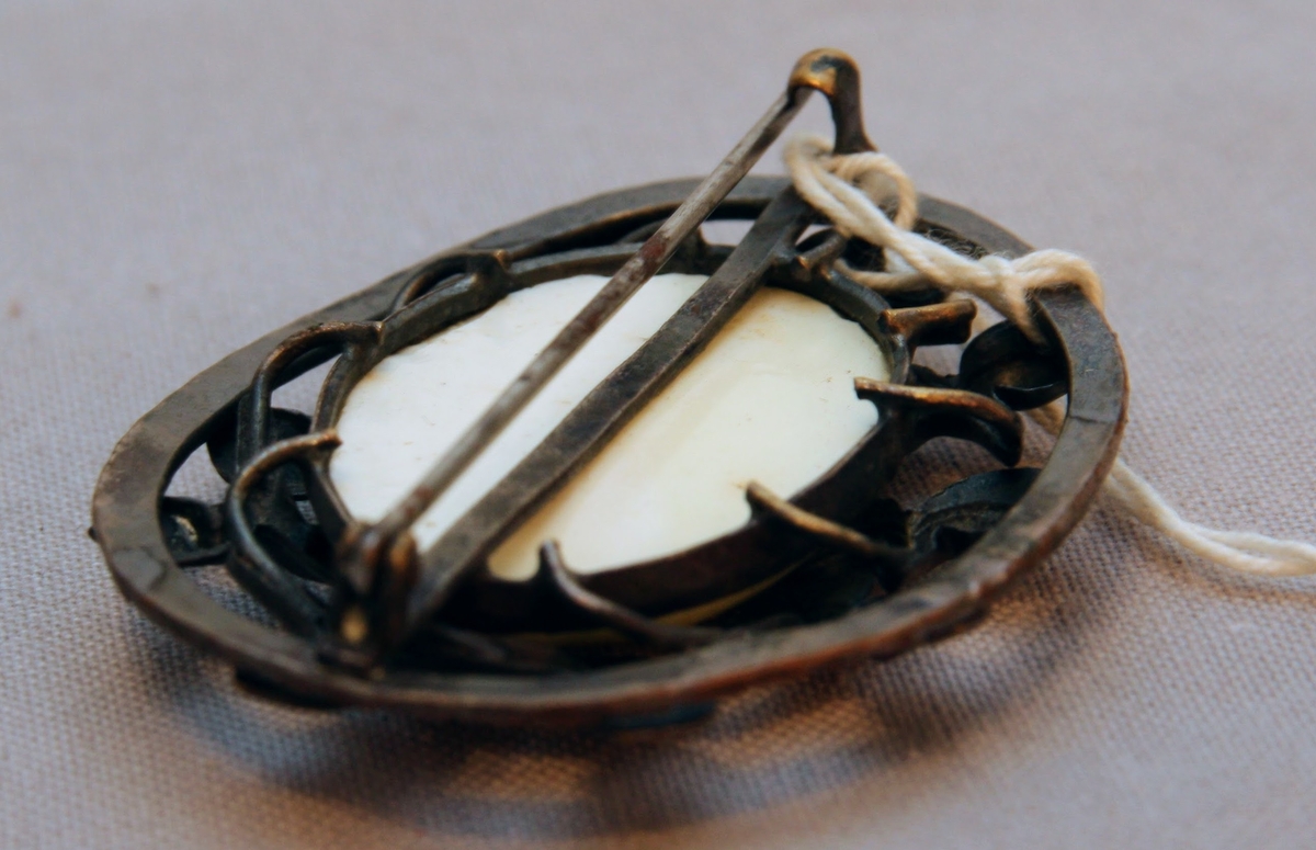 Brosch med en oval uppåtbuktad och polerad benplatta omkransad av en ring stiliserade bladmönster av försilvrad koppar.