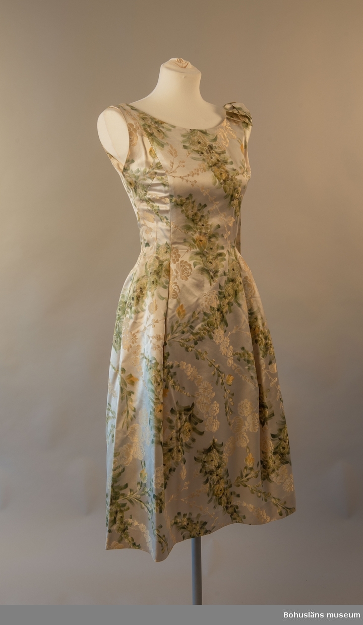Ärmlös och A-linjeformad klänning med gula och gröna blom- och växtornament mot ljusgul botten. På vänster axel ett band fäst som hänger ned på baksidan. Dragkedja bak som slutar mellan skuldrorna.

Underkjol fäst i midja i kraftigare syntetmaterial som hjälper till att skapa kjolens fall.