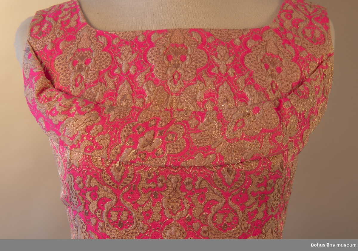 Klänning med beigt och guldfärgat mönster mot rosa botten. Rosett baktill som för blicken fäster det draperade tyget mellan skuldrorna. Framsida utsmyckad med en bård över bysten.