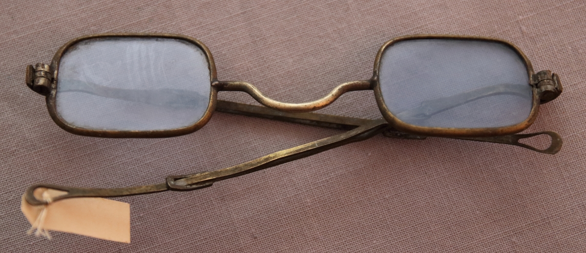 Glasögon med mässingsomramning och förlängbara skalmar av samma metall. Ljusblå glas av rektangelform med avrundade hörn. Med utfällbara skalmar som avslutas i öglor.