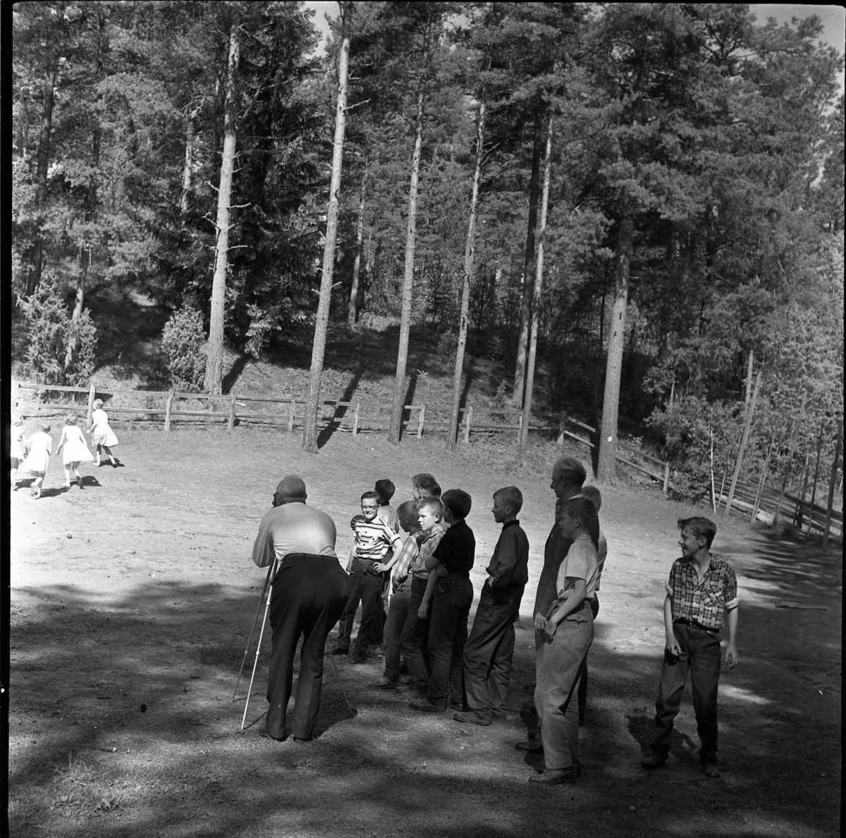 Barn leker på en gräsmatta vid ett skogsbryn. Några pojkar står bredvid och tittar på. En man står och fotograferar de lekande barnen.