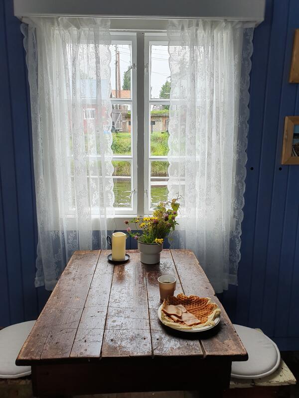 Foto av interiør i hvilebrakke for overnatting. Bord foran vindu, blomster og tallerken med vaffel på bordet.