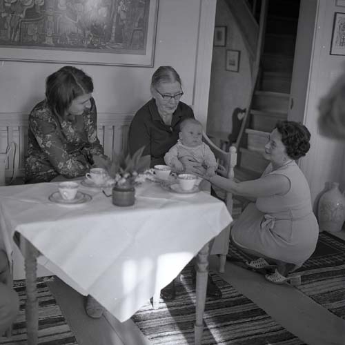Lill-Annas dop sommaren 1973, här i ett rum på gården Sunnanåker. Två kvinnor sitter vid ett dukat kaffebord och den äldre av dem håller dopbarnet i knät. Adéle sitter på huk bredvid dem.