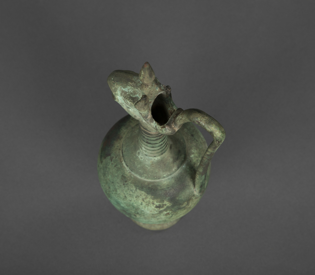 Skulptur av metall. Med en liten stett, hank og fantasidyr hode (fugl/enhjørning). Omtalt som vase men har en veldig grunn åpning i fantasidyrets hode og ut gjennom munnen. Irrgrønn.