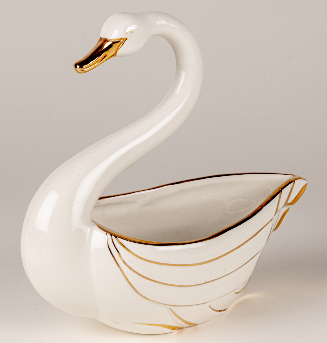 Prydnadsfigur eller skål i form av en svan, flintgods, modell A, dekorserie Aladdin; vit glasyr med handmålat guld. Aladdin producerades under åren 1941-1943. Oklart vem formgivaren är.