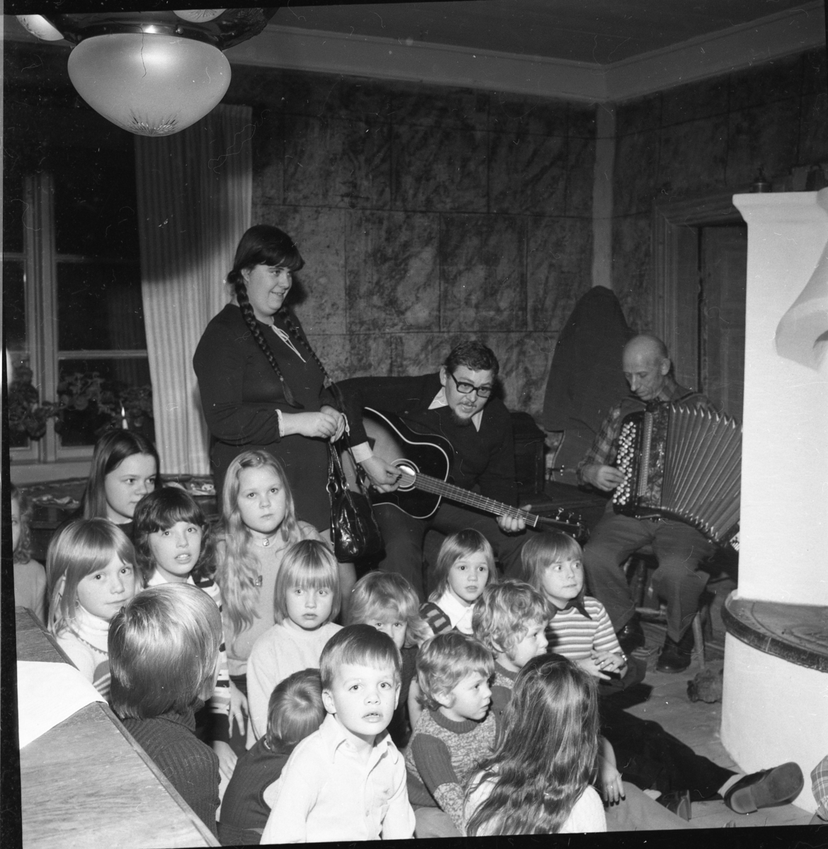 Vuxna och barn i en stuga med öppen spis. Två män spelar gitarr respektive dragspel. Det är Ölmstad hembygdsförenings julfest.