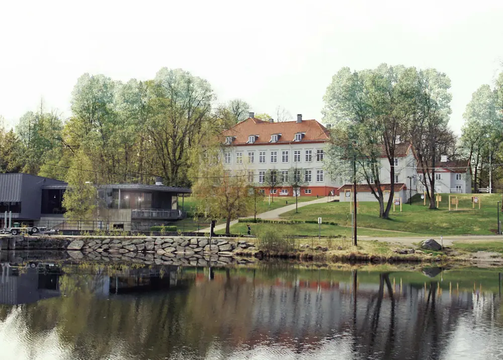 Foto av nordlige sidefasade av Eidsvollsbygningen delvis skjult bak grønne trær. I forgrunnen renner Andelva rolig forbi, bygningen og trærne speiles i vannet.