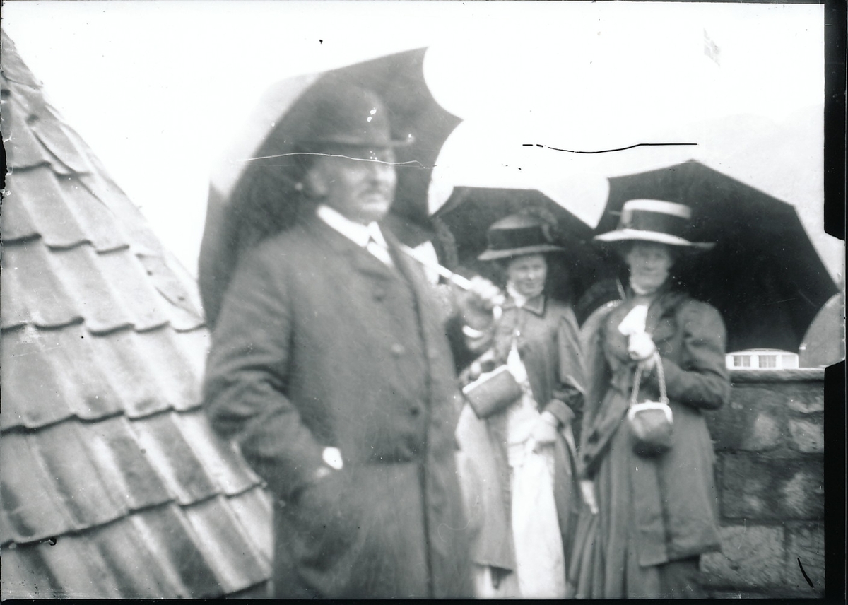 Bestyrer Wilhelm Rydgren, Signe Rydgren og Nicoline Rydgren med paraplyer, står ved bygning med tak av teglsstein som går helt ned til bakken. En mur i bakgrunnen.