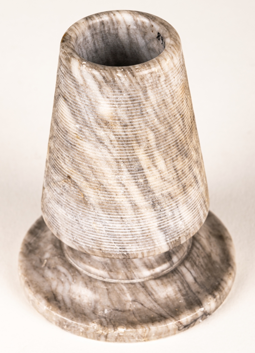 Tändsticksställ, av gråvit marmor, i form av stympad kon på rund fot. Hållaren ovanpå räfflad.