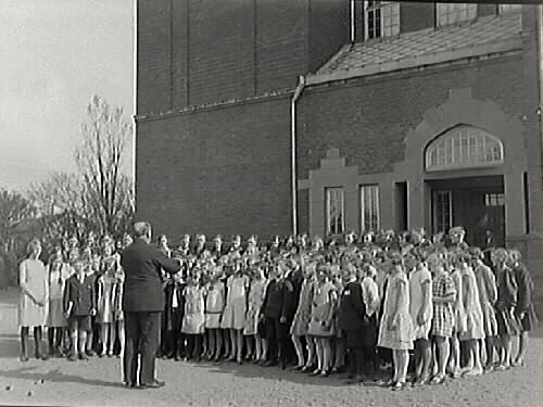 Barnkör sjunger utanför Rosenfredsskolan (Södra folkskolan). En man dirigerar kören.