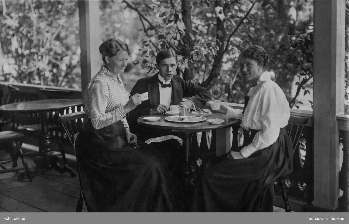 Två kvinnor och en ung man sitter vid ett kaffebord på verandan till utvärdshuset Tivoli. På bildens baksida står: Från Tivoliverandan husmor, kassörskan och en biljardgrabb. Ur en samling som tillhört Skoglund från Långskog, Matfors.