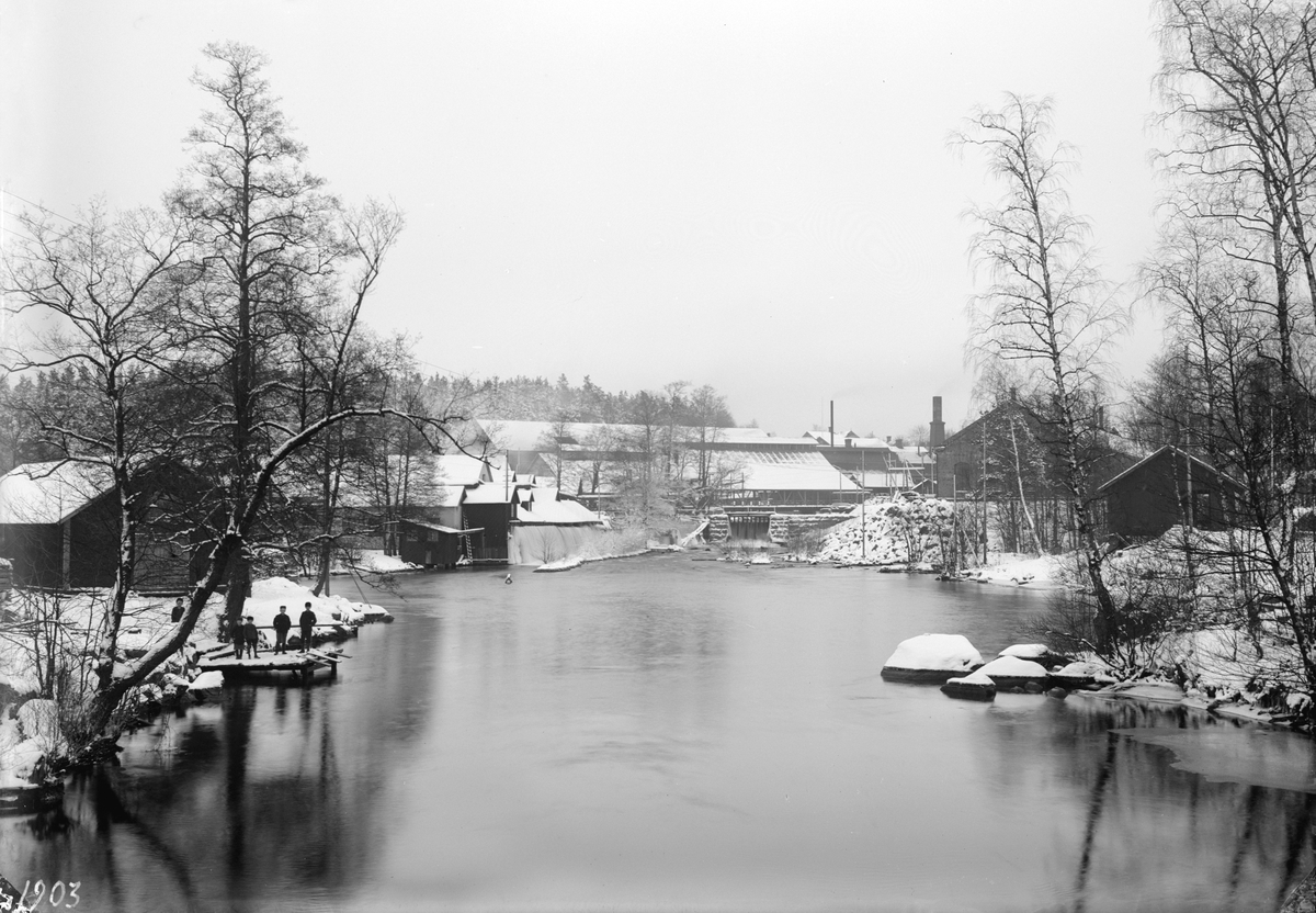 Vinterbild från Boxholm 1903. I fonden ses följande anläggningar vid Boxholms bruk. Från vänster, klippspiksfabriken, Nedre verket (grovverket) och Nya mekaniska verkstaden. De sistnämnda byggnaderna var vid tiden för bilden nyuppförda. Vy från Bredgårdsbron.