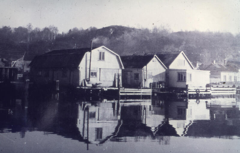 Sort-hvitt-bilde som viser en rekke hvite bygninger som ligger helt nede i vannkanten. Vannflaten er speilblank. (Foto/Photo)