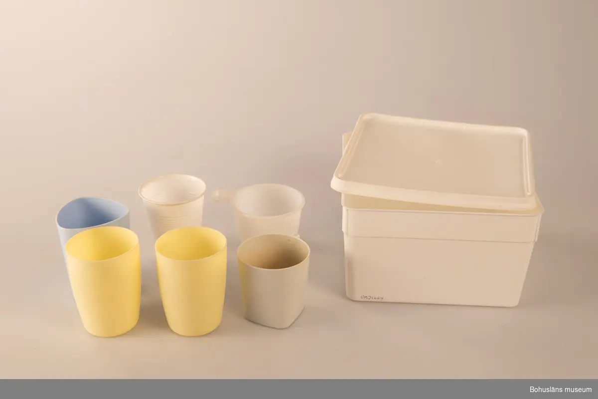 Kvadratisk låda av vit plast med plastlock. I lådan förvaras sex muggar/koppar: två gula plastmuggar, en ljusblå plastmugg, en vit plastmugg med handtag,  en engångsmugg av plast och en engångsmugg med handtag av plast.