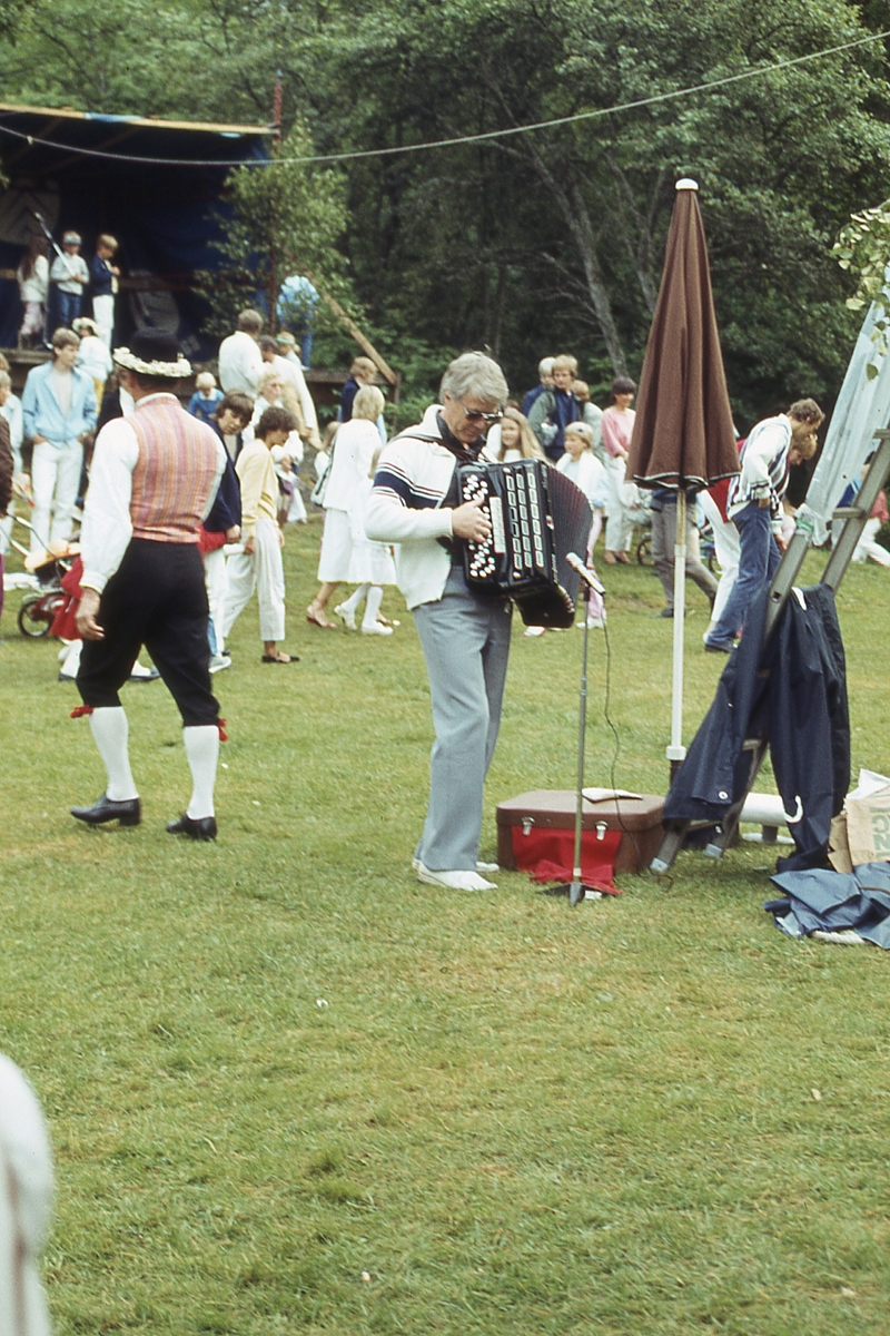 Barn och vuxna dansar kring stången vid midsommarfirande på Ekensås cirka 1982. En regnig aftor där spelmannen Frank Efraimsson spelar dragspel.