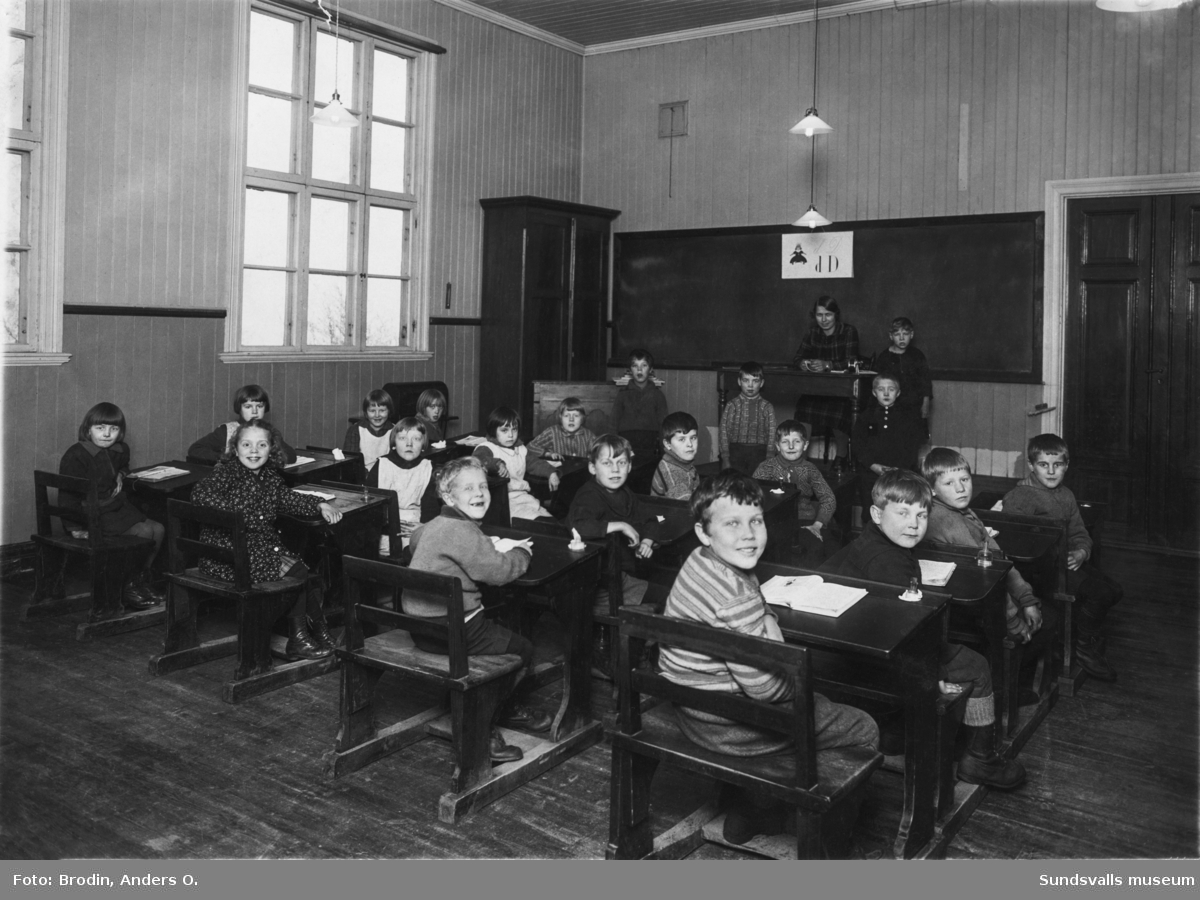 Skolfoto i ett klassrum med barnen vid sina bänkar och lärarinna vid katedern. Ur en samling som tillhört Allan Hallén, Essvik.