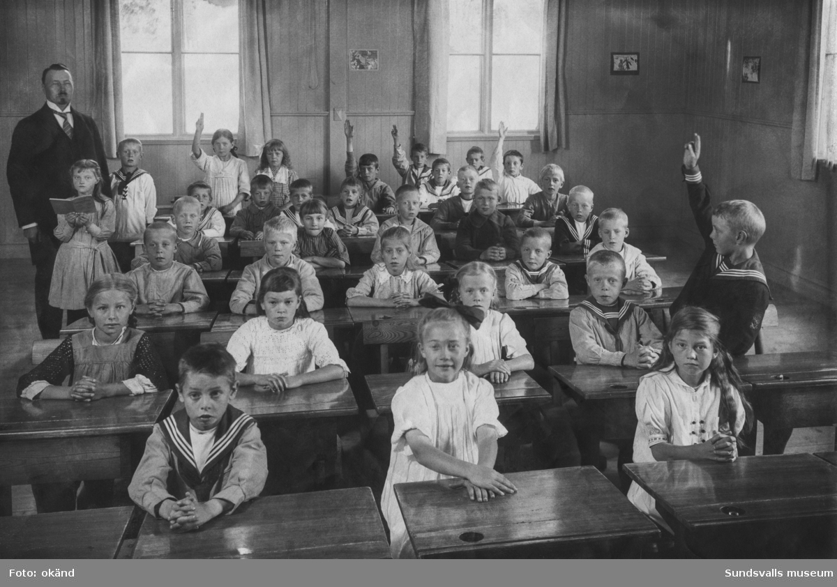 Skolfoto, barnen sitter vid sina bänkar i ett klassrum med träpanelade väggar. Manlig lärare. Troligen någon skola i Timrå eller Sundsvall.