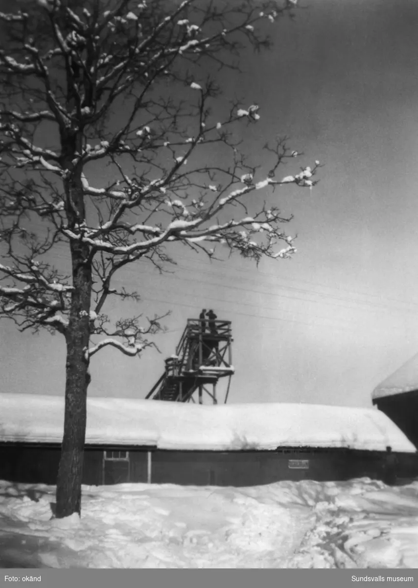 Kyrkstallarna vid Sättna kyrka i vinterskrud. Bakom syns tornet till hoppbacken som fanns vid Ånäsvallen, två personer syns stå uppe i tornet. På bildens baksida står: Kyrkstallarna Vallen. Skidbacken i bakgrunden, 1940-talet. Ur en samling från Byn, Norrbäcken, Sättna.