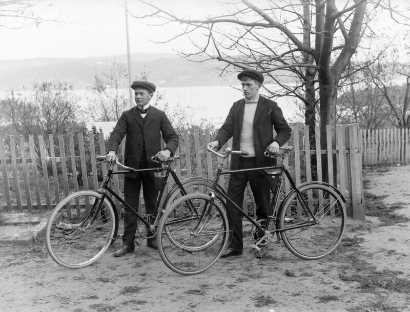 Sort-hvitt-bildet viser to gutter i dress og luer med hver sin sykkel. De står i en hage. I bakgrunnen skimtes fjorden. (Foto/Photo)