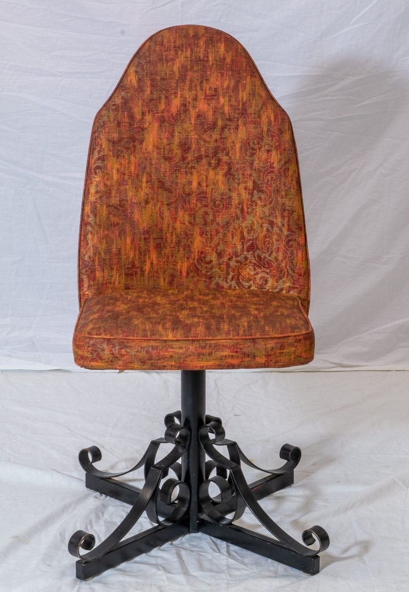 Spisestuestol med rødbrunt trekk. Bein i sort metall minner om smijern og bakplate i plast med tremønster. Stolen kan snurre.
