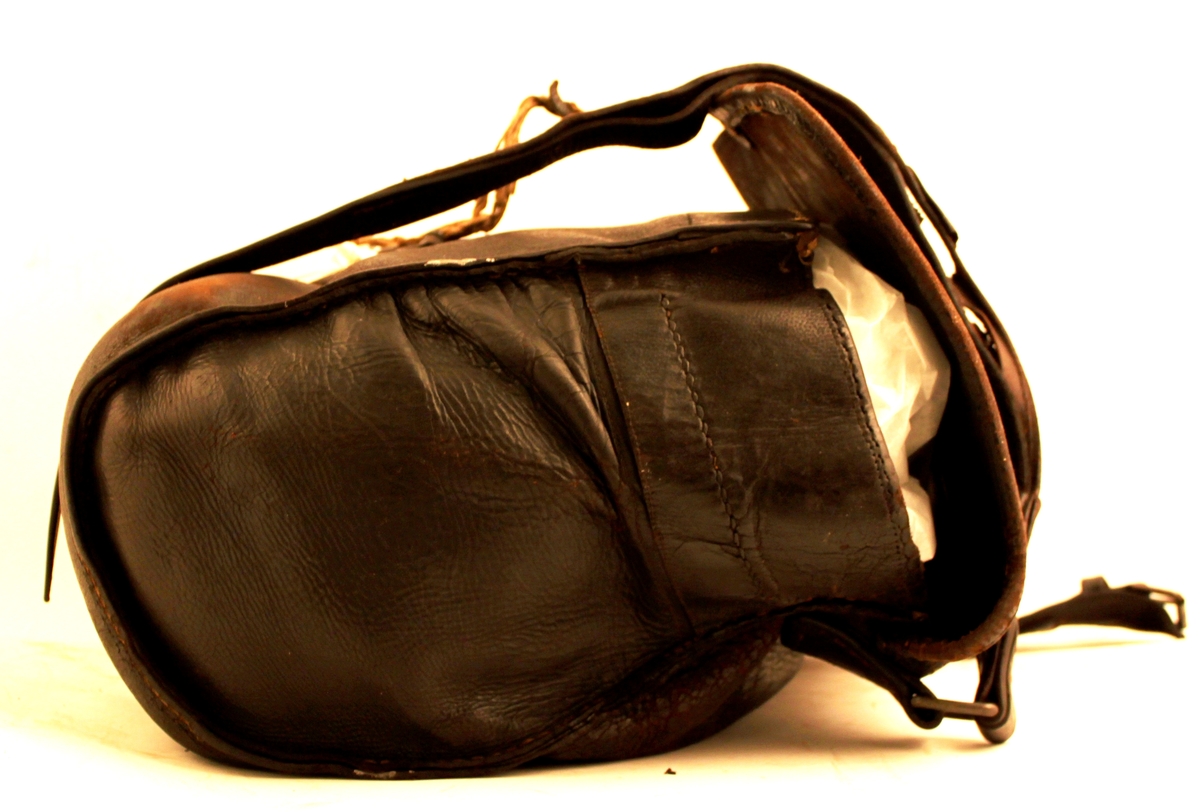 Brevbärarväska av svärtat läder med bärrem och spänne. Inuti väskan på baksidan två mindre fack. 

Ett knippe snören fastbunda vid spännet. Hjälp för att stänga väskan?

Lädret slitet.