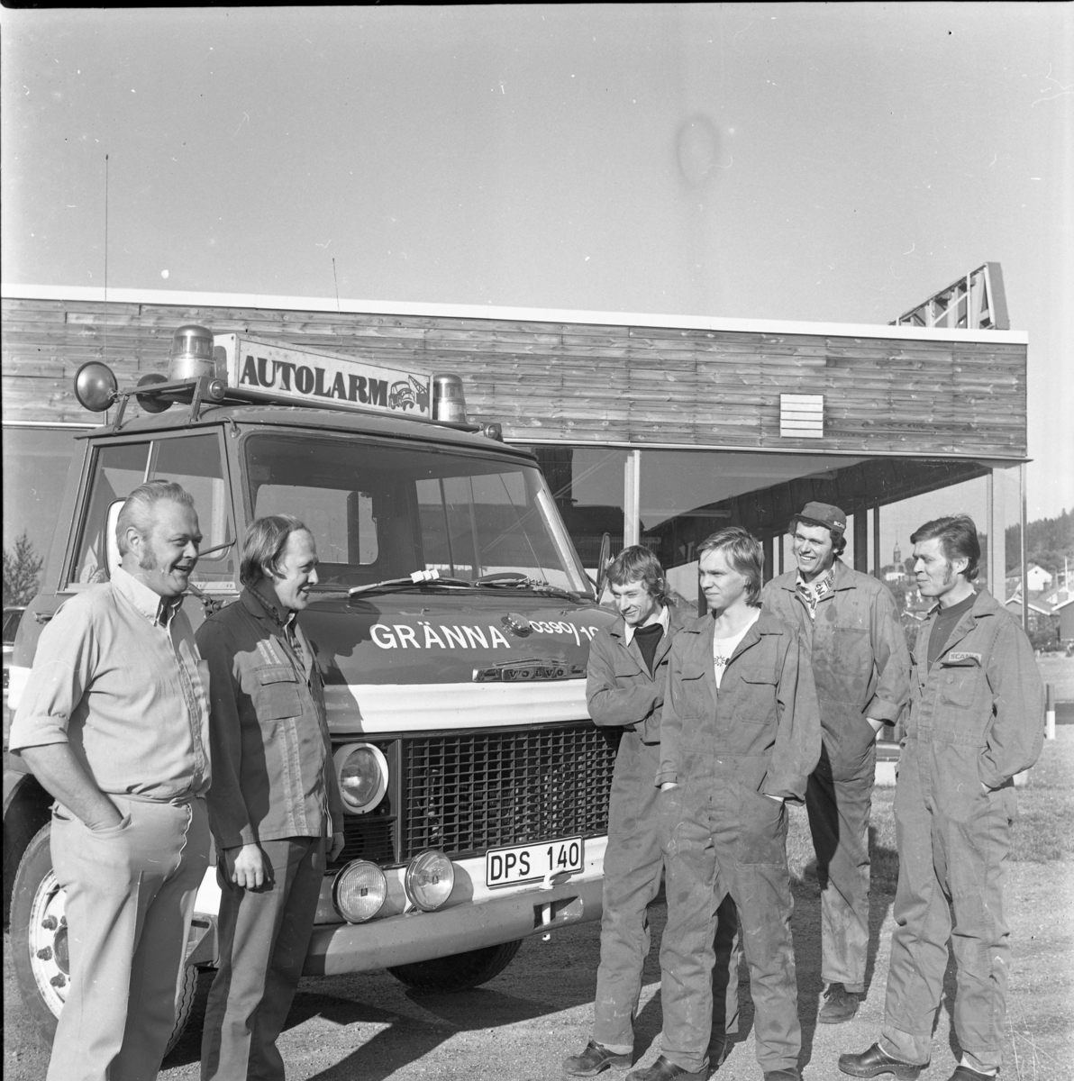 Sex män står vid fronten till en bärgningsbil med varningslampor och skylt "Autolarm" på taket. Två av männen, i civila kläder och något äldre, står till vänster. De andra fyra bär verkstadsoverall.