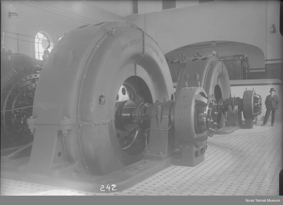 Maskinsalen på Rygene kraftstasjon med generatorer og turbin, en funksjonær i bildets bakgrunn; turbinene som er levert av Myrens Verksted er dobbelte, horisontale francisturbiner med en samlet ytelse på 9000 hk ved 21 m fallhøyde, 250 o/m og vannforbruk ved full last 28 m3/s : kraftstasjonen eies av A/S Rygene Tremassefabrikker og ble bygget i 1913-14, senere ble det bygget ytterligere to kraftstasjoner tilhørende samme anlegg