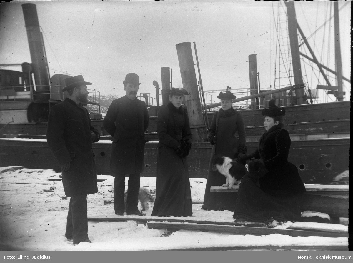 Kvinner og menn i reiseantrekk på kaien med dampskip i bakgrunnen