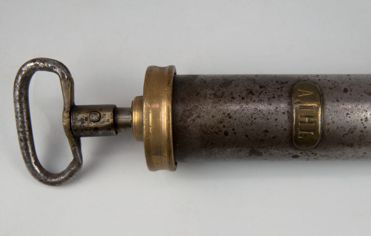 Oljespruta av metall. Cylinderformad men lång krökt pip längst fram. Ovalt handtag fäst i locket. På sprutan sitter en mässingsskylt med texten "A. IHL".