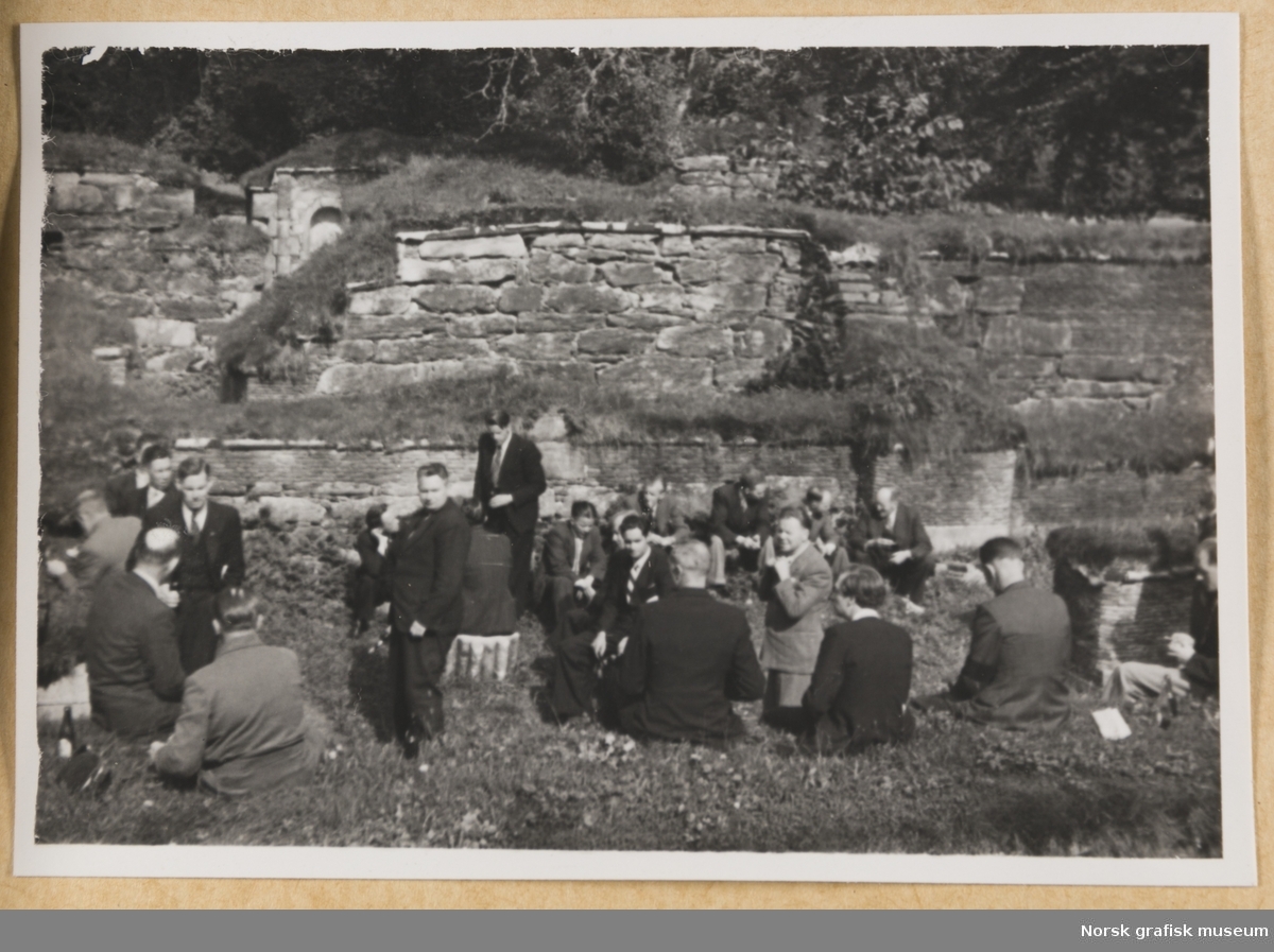 Utendørsmotiv. En gruppe menn i dress og slips sitter og står rundt i et ruin- og naturlandskap. Fra Lysekloster. Fotografert i forbindelse med Vestlandsk Trykkerstevne i Bergen, 1946.