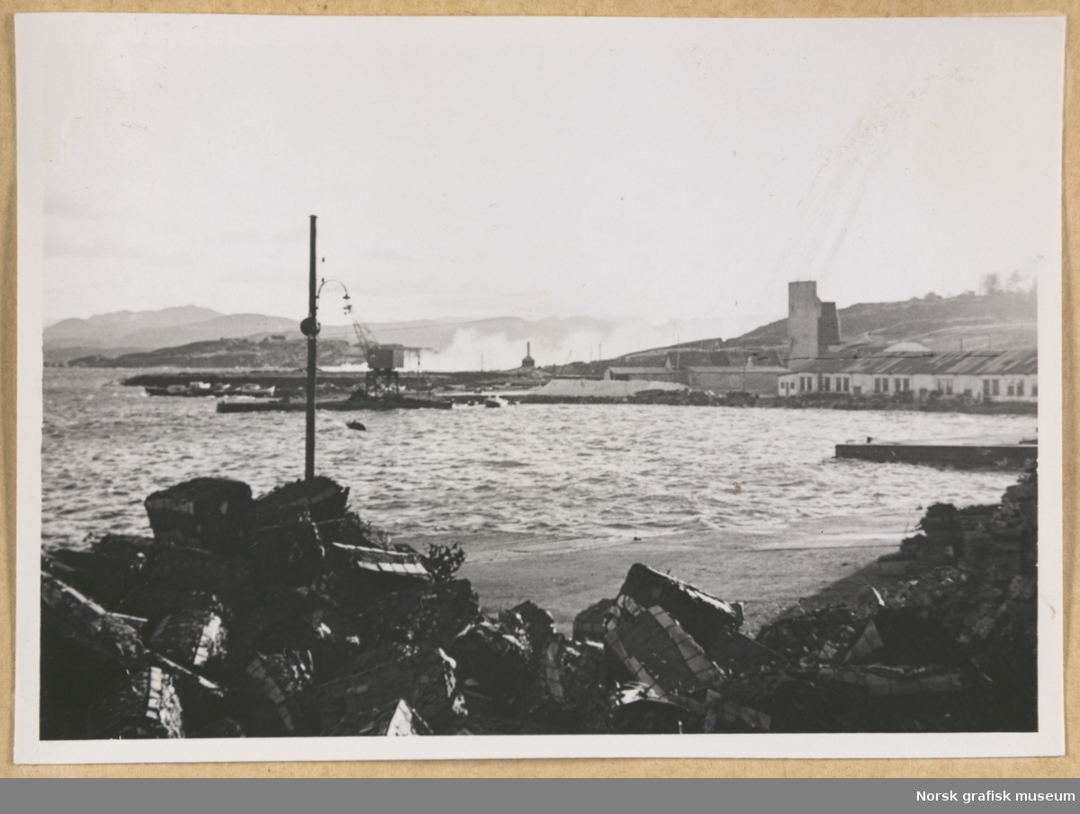 Kystlandskap. I forgrunnen en strand med hauger av vrakgods eller fiskeutstyr. I mellomgrunnen moloer og lave lagerbygninger ved sjøen. I bakgrunnen øyer eller lave åser. Dette kan muligens være i Breivik på Storhaug, sett mot Marøy. Fotografert i forbindelse med Vestlandsk Trykkerstevne i Stavanger, 1948.