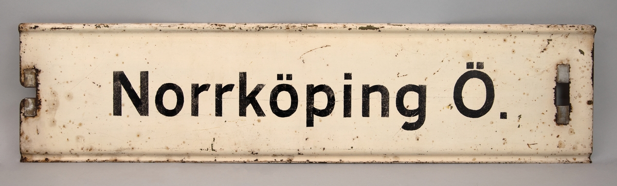 Rektangulär destinationsskylt av målad plåt. Vit botten med svart text, på ena sidan står det "Norrköping Ö.", på andra sidan står det "Finspång". Hål på kortsidorna för upphängning.
