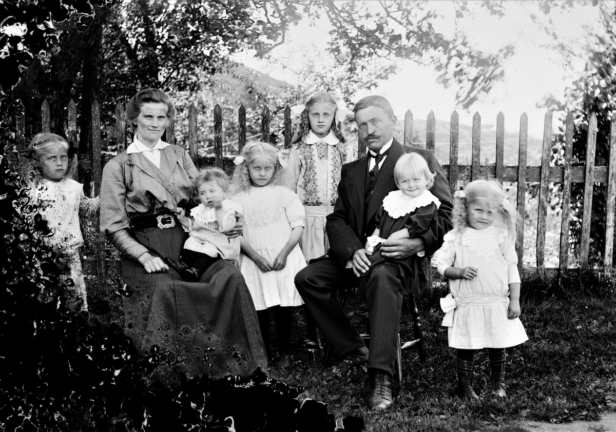 Makarna Hilma och Vilhelm Andersson sitter med sina barn vid ett spjälstaket, Dalhem Grimmared. Från vänster räknat heter barnen Anna, Margareta, Elin, Helfrid, Märta och Signe.
(Se även bild nr GEA047 och GEA076)