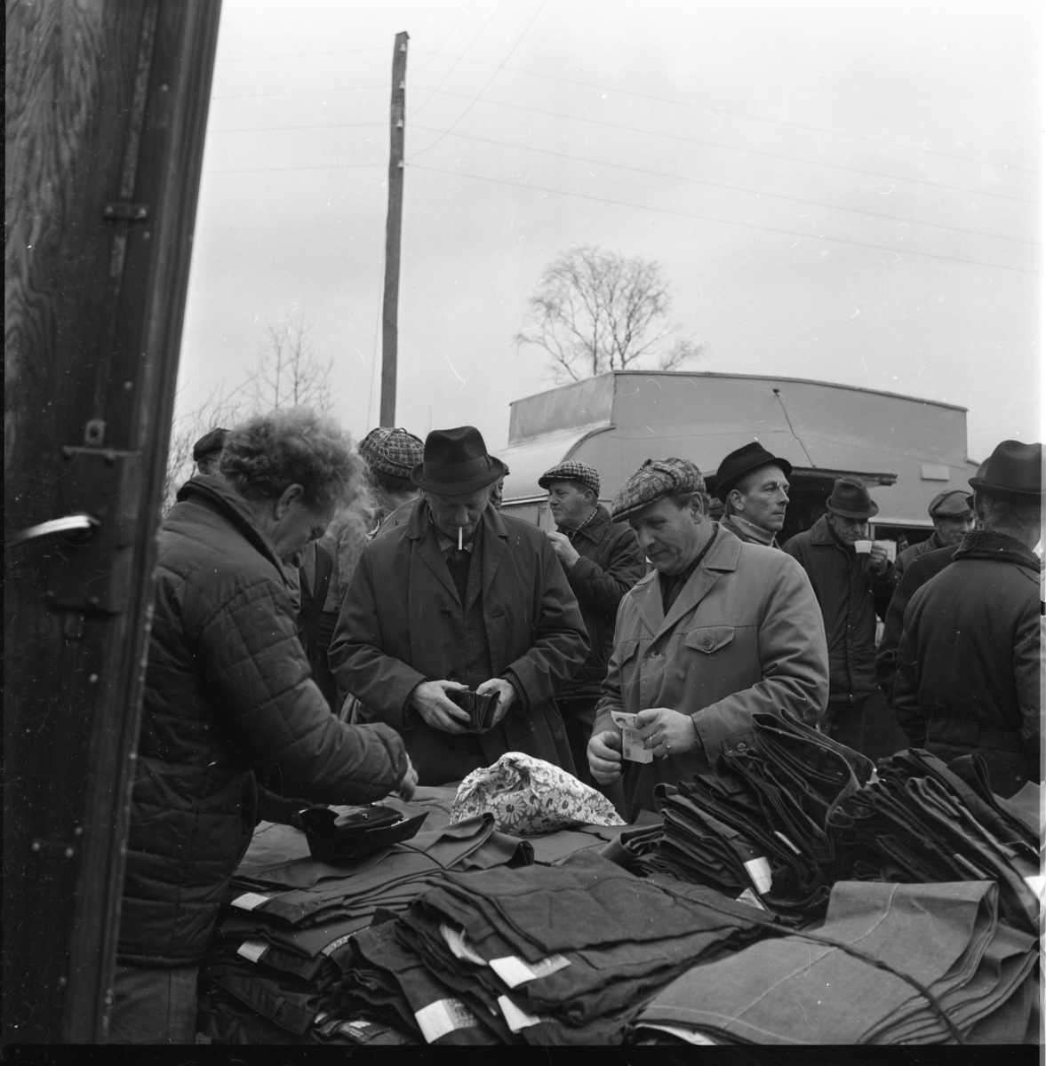 Ett par män med pengar och börs i handen står vid ett bord med ett stort antal byxor. Försäljning i anslutning till auktion på Vireda gård i Lekeryd.
