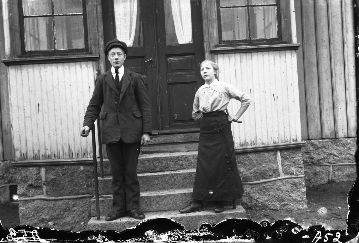 En ung man i kostym och en yngre flicka står på verandatrappan till småskolan i Sundbo, Sällstorp. (Negativet skadat nedtill. Se även bild på ynglingen nr GEA033)