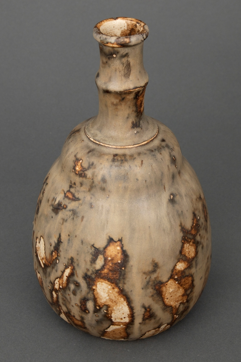 Vase i glasert steingods. Vasen har pæreform med lang smal hals og ringformet munningsrand. Store deler av yttersiden er dekket av en gråfarget flyteglasur, men tidvis kommer brune og uglaserte partier til syne.