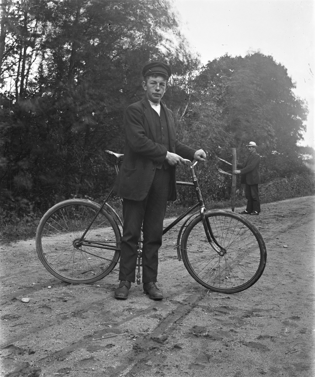 En ung man vid namn Hansson från Hansa, Syllinge i Veddige står tvärs över grusvägen med en cykel. I bakgrunden står en annan man på vägen med en spetsad stolpe. (Se även bild nr GEA028)