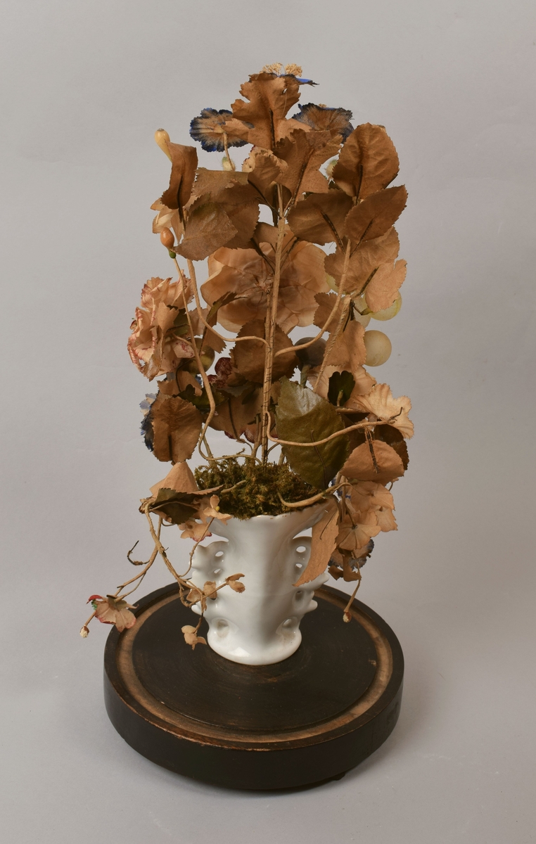 Oppsats med kunstige blomster og frukt i liten porselensvase. Vasen med bladmotiv og forgylling på forsiden. Hele oppsatsen dekket med en glasskuppel.